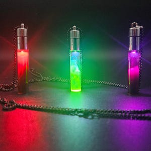 Puede incluir: Tres collares con colgantes iluminados con viales cilíndricos de vidrio. Los viales están llenos de líquidos de colores, que brillan en rojo, verde y morado. Cada colgante cuelga de una cadena negra. El fondo es oscuro.