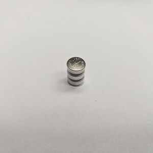 K&ouml;nnte beinhalten: Ein Stapel von vier kleinen, silberfarbenen Knopfzellenbatterien. Die oberste Batterie hat ge&auml;tzte Markierungen. Die Batterien sind auf einer wei&szlig;en Oberfl&auml;che gestapelt.