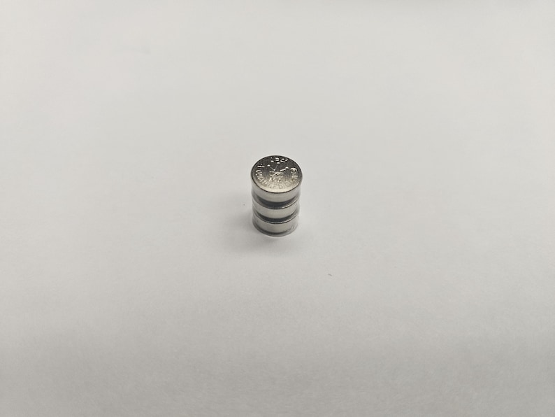 K&ouml;nnte beinhalten: Ein Stapel von vier kleinen, silberfarbenen Knopfzellenbatterien. Die oberste Batterie hat ge&auml;tzte Markierungen. Die Batterien sind auf einer wei&szlig;en Oberfl&auml;che gestapelt.