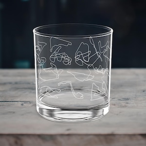 Könnte beinhalten: Ein klares Whiskyglas mit weißem Umrissdesign verschiedener Rennstrecken. Das Glas steht auf einer hellen Holzoberfläche mit verschwommenem dunklen Hintergrund. Das Glas ist zylindrisch mit einem dicken Boden.
