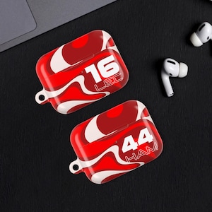 May include: Two red and white patterned AirPod cases, each with a different number and name printed on them. One case has "16 LEC" and the other has "44 HAM". Two white AirPod earbuds are also visible. The cases are rectangular with rounded edges.