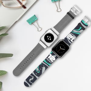 Könnte beinhalten: Zwei Apple Watches mit verschiedenen Armbändern. Eine Uhr hat ein graues Lederarmband, die andere ein marineblaues Armband mit einem türkis-weißen Muster und dem Text "12 ANT". Die Uhren liegen auf einer weißen Oberfläche.