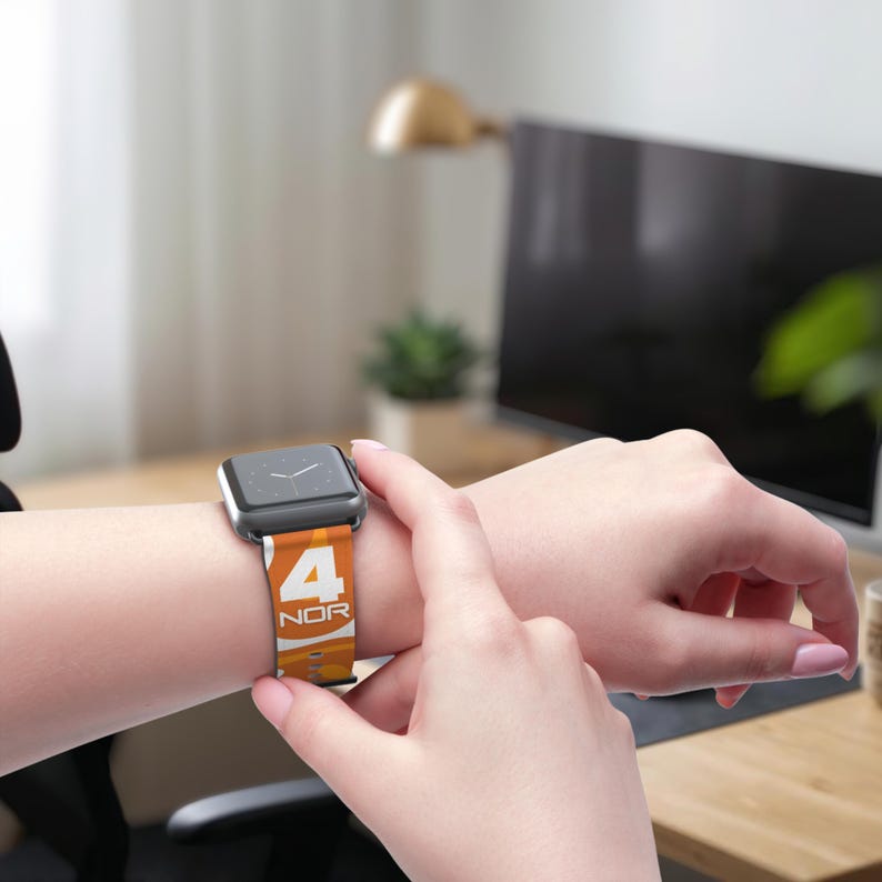 May include: An orange and white watch band with the number 4 and the letters NOR printed on it. The watch band is attached to a silver smartwatch. The watch is on a person's wrist.