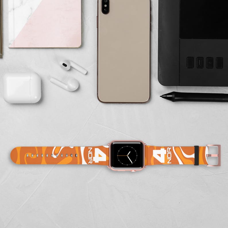 May include: An orange and white patterned Apple Watch band with the number 4 and the letters NOR printed on it. The watch face is black with a rose gold case. Other items on the table include a phone, earbuds, and a notepad.