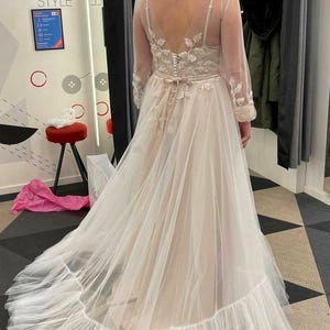 May include: A white tulle wedding dress with a lace bodice and long sleeves. The dress has a flowing skirt with a ruffled hem.