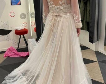 May include: A white tulle wedding dress with a lace bodice and long sleeves. The dress has a flowing skirt with a ruffled hem.