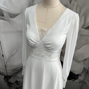 Könnte beinhalten: Ein weißes, langärmeliges Brautkleid mit V-Ausschnitt und einem Spitzenoberteil. Das Kleid hat ein tailliertes Oberteil und einen fließenden Rock.