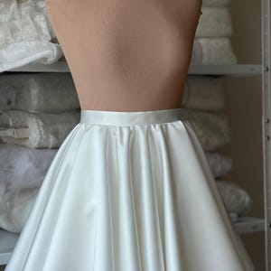 May include: A white satin skirt with a wide waistband is displayed on a mannequin. The skirt has a flared A-line shape, with soft folds and a smooth, shiny texture. The background shows shelves with fabric.
