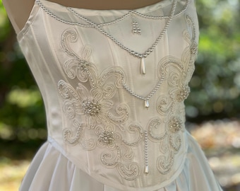 May include: A white satin wedding dress with a strapless bodice. The bodice is embellished with intricate silver beading and embroidery, including floral and swirl designs. The skirt is full and flowing, creating a classic bridal look.