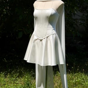 May include: A white strapless mini dress featuring a structured corset-style bodice with vertical seams and a flared skirt. The garment includes a long, flowing sheer white cape or scarf draped over the shoulders that extends down the back.