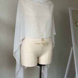 May include: A sheer white chiffon shawl draped over a beige dress form. The fabric is lightweight and translucent, falling in soft, asymmetrical folds around the torso. The dress form features the text Royal Dress forms Italy design.