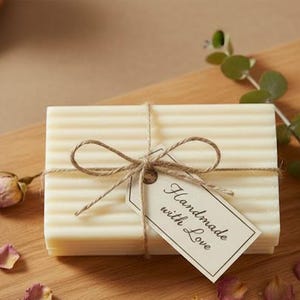 May include: A rectangular bar of light-colored soap, wrapped with twine and a tag that reads "Handmade with Love." The soap is on a wooden cutting board, decorated with a sprig of eucalyptus and rose petals. Other soaps are in the background.