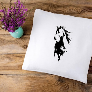 May include: White square pillow with a black horse silhouette design. The horse is depicted in a running pose with its mane flowing. The pillow is on a wooden surface, with a small turquoise vase of purple flowers in the background.