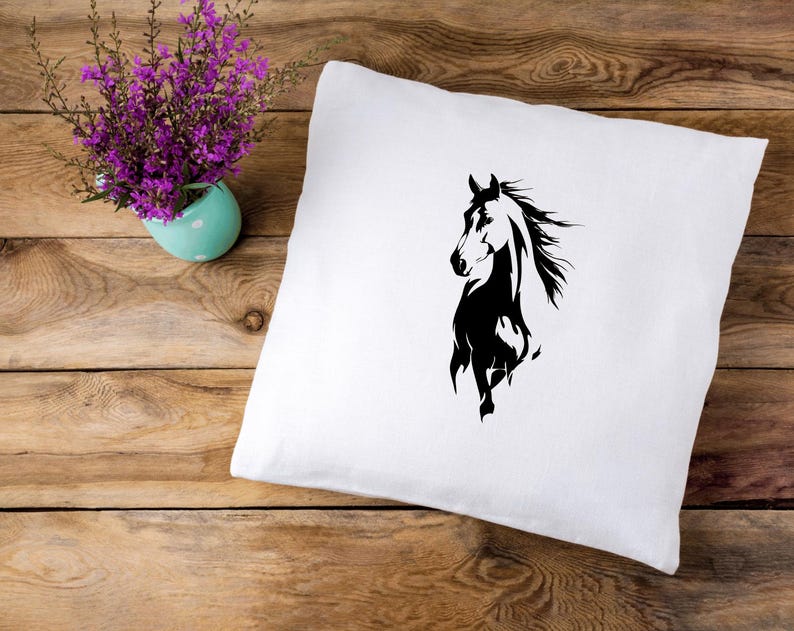 May include: White square pillow with a black horse silhouette design. The horse is depicted in a running pose with its mane flowing. The pillow is on a wooden surface, with a small turquoise vase of purple flowers in the background.