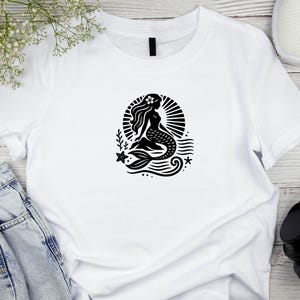 May include: White t-shirt with a black silhouette of a mermaid sitting on a rock with a star and waves behind her.
