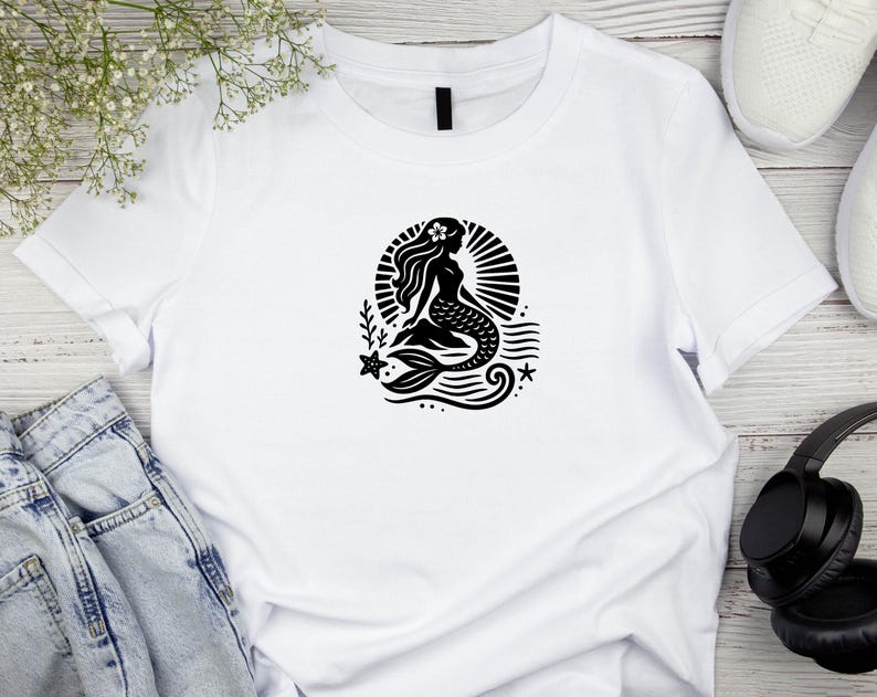 May include: White t-shirt with a black silhouette of a mermaid sitting on a rock with a star and waves behind her.