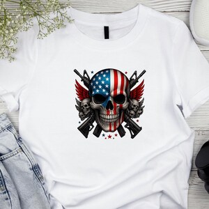 May include: White t-shirt featuring a skull design with an American flag motif. The skull has a blue and white stars and stripes pattern, with red wings and two rifles crossed behind it. The design includes red stars.