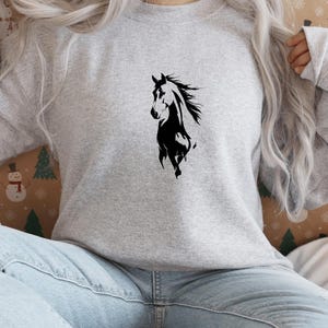 May include: A light gray sweatshirt featuring a black silhouette of a horse. The horse is depicted in a dynamic pose, with its mane flowing. The sweatshirt is made of a soft material and is suitable for casual wear.