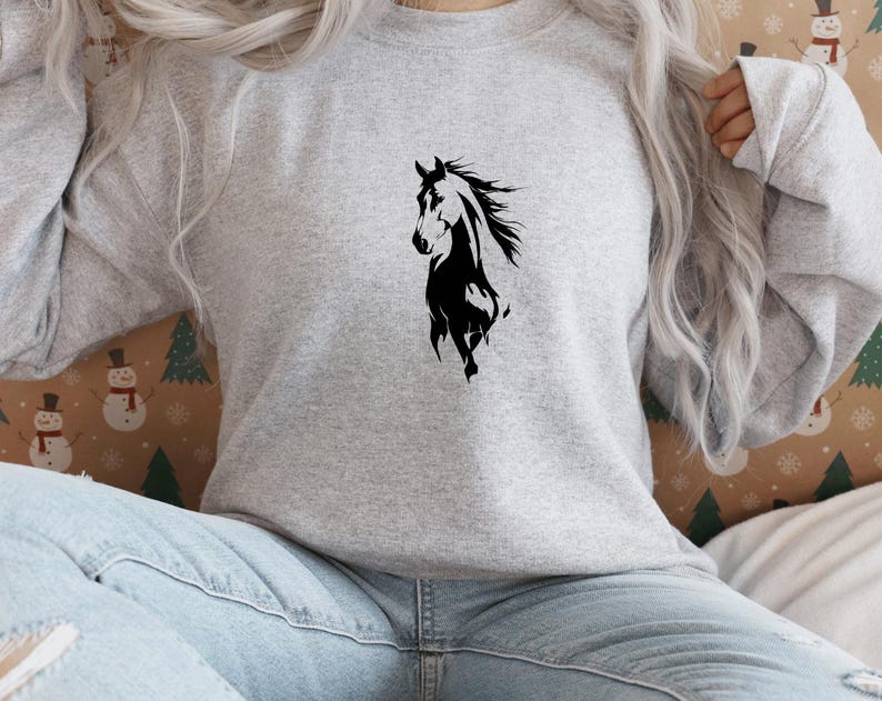 May include: A light gray sweatshirt featuring a black silhouette of a horse. The horse is depicted in a dynamic pose, with its mane flowing. The sweatshirt is made of a soft material and is suitable for casual wear.