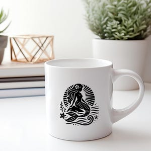 May include: White ceramic mug with a black silhouette of a mermaid with a flower in her hair, sitting on a wave with a starfish and a seashell. The mermaid is surrounded by a sunburst.