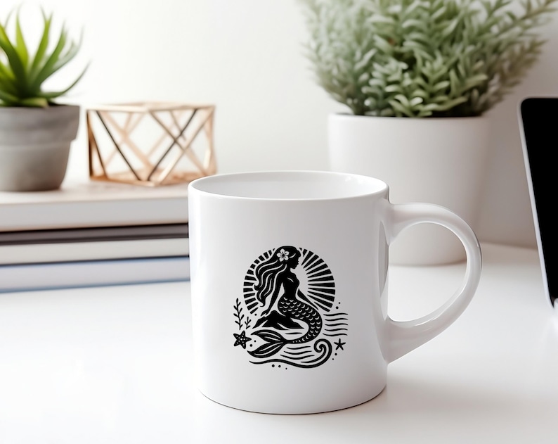 May include: White ceramic mug with a black silhouette of a mermaid with a flower in her hair, sitting on a wave with a starfish and a seashell. The mermaid is surrounded by a sunburst.