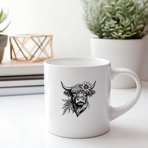 May include: White ceramic mug with a black and white illustration of a Highland cow with a flower in its hair. The cow is looking to the right.