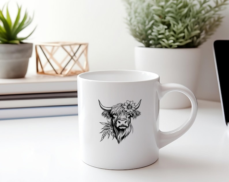 May include: White ceramic mug with a black and white illustration of a Highland cow with a flower in its hair. The cow is looking to the right.