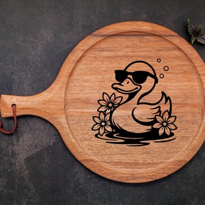 May include: A wooden serving board with a handle, featuring a black and white illustration of a duck wearing sunglasses and surrounded by flowers.