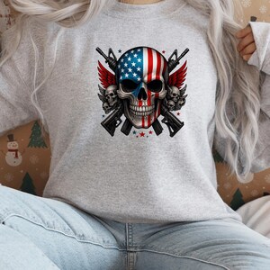 May include: A light gray sweatshirt featuring a skull design with an American flag motif. The skull has red and white stripes and blue stars. Two rifles are crossed behind the skull, with red wings and smaller skulls on either side.