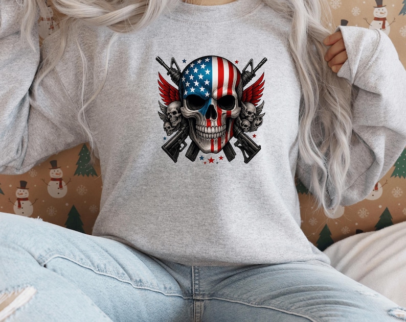May include: A light gray sweatshirt featuring a skull design with an American flag motif. The skull has red and white stripes and blue stars. Two rifles are crossed behind the skull, with red wings and smaller skulls on either side.