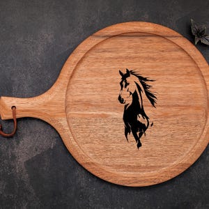 May include: A round wooden serving tray with a handle and a leather loop. The tray features a black silhouette of a horse's head and flowing mane. The wood has a natural grain and a warm, light brown color.