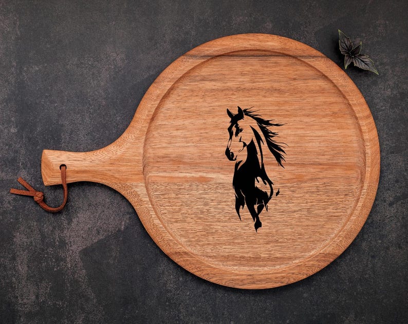 May include: A round wooden serving tray with a handle and a leather loop. The tray features a black silhouette of a horse's head and flowing mane. The wood has a natural grain and a warm, light brown color.