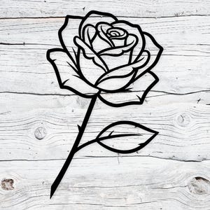 May include: A black outline of a rose, featuring detailed petals and a stem with a single leaf. The design is cut from a single piece of material. The rose is set against a weathered, white wooden background.