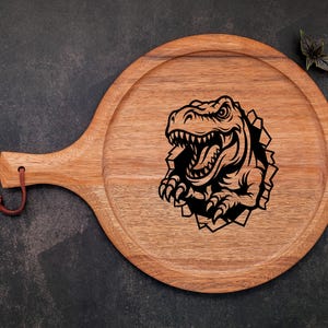 May include: A wooden serving board with a handle, featuring a black and white illustration of a dinosaur head breaking through the surface.