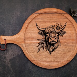 May include: A wooden serving board with a handle, featuring a black and white engraved illustration of a Highland cow with flowers in its hair.