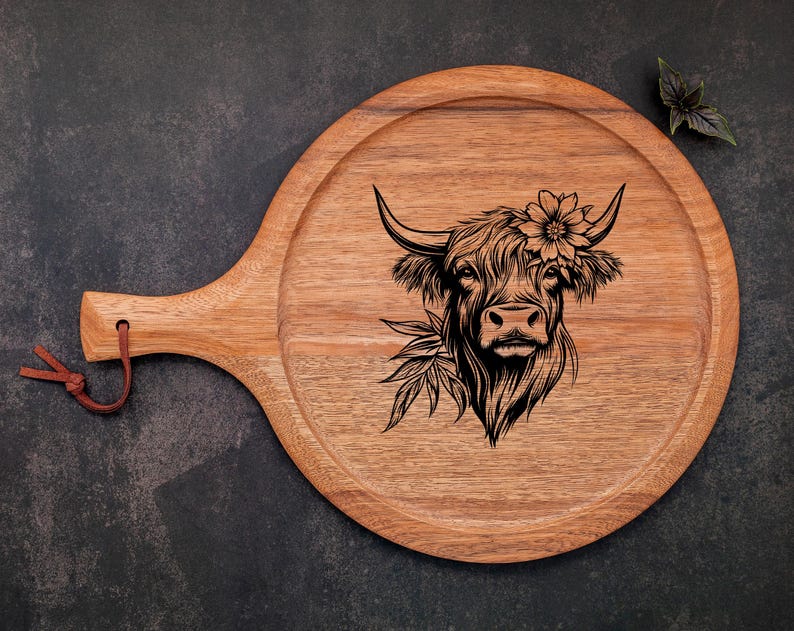 May include: A wooden serving board with a handle, featuring a black and white engraved illustration of a Highland cow with flowers in its hair.