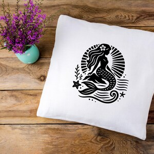 May include: A white pillow with a black silhouette of a mermaid with a flower in her hair. The mermaid is sitting on a wave with a starfish and a seashell.