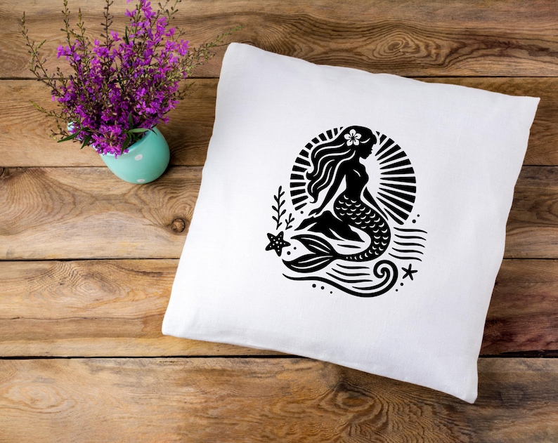 May include: A white pillow with a black silhouette of a mermaid with a flower in her hair. The mermaid is sitting on a wave with a starfish and a seashell.