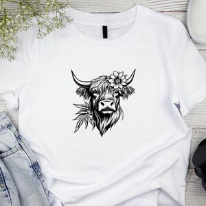 May include: White t-shirt with a black and white illustration of a Highland cow with a flower crown.