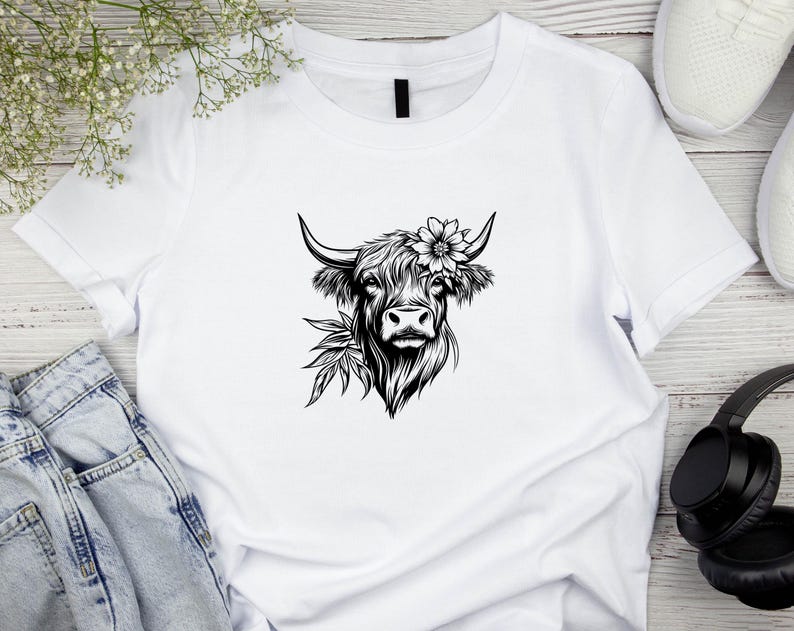 May include: White t-shirt with a black and white illustration of a Highland cow with a flower crown.