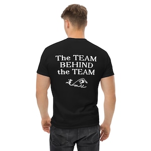May include: Black t-shirt featuring the white text "The TEAM BEHIND the TEAM" and a swimmer graphic. The shirt is made of a soft material. The design is centered on the back of the shirt, suitable for casual wear.