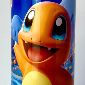 Puede incluir: Un vaso cilíndrico con un personaje de dibujos animados Charmander en naranja y amarillo, sobre un fondo azul, con una pajita plateada. El diseño incluye estrellas y relámpagos.
