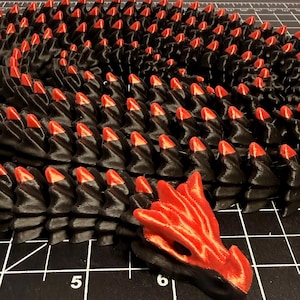 May include: A 3D-printed articulated dragon figure featuring a black body with red accents on the scales and a solid red head. The dragon is coiled on a black cutting mat with white grid lines and visible inch markings, including the numbers 5 and 6.