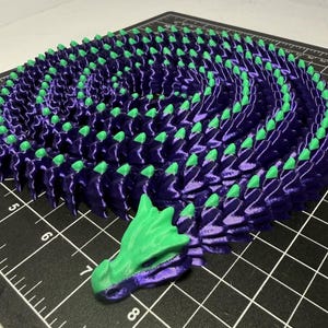 May include: A 3D printed articulated dragon figure coiled in a spiral shape. The dragon features a metallic purple body with bright green accents on the head and along the spine. It is positioned on a black cutting mat with white grid lines.