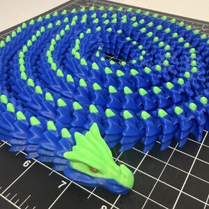 May include: A 3D-printed articulated dragon toy in blue and green. The dragon is coiled, with blue scales and green accents on the spine and head. The dragon's head is at the end of the coil.