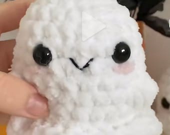 May include: A fluffy, white crochet ghost with black button eyes and a stitched smile. The ghost has pink cheeks and a soft texture. This Halloween-themed plush toy is ideal for seasonal decor or as a gift.