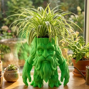 Op de afbeelding: Een groene, grillige plantenbak in de vorm van een bebaarde man met armen en benen. De plantenbak bevat een levendige groen-witte graslelie. De plantenbak is gemaakt van een stevig materiaal en staat op een houten oppervlak bij een raam.