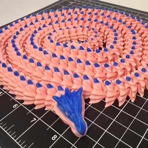 May include: A pink and blue dragon sculpture. The body is a spiral of pink, textured segments, each tipped with a blue point. The dragon's head is blue, with detailed features.