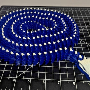 May include: A long, articulated dragon figure coiled in a spiral shape. The body is deep blue with small, white, triangular spikes running along the spine. The head is white with blue accents, and the entire figure rests on a black cutting mat.