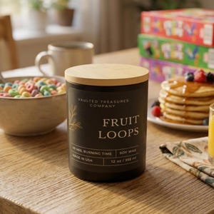 May include: A black candle with a wooden lid, labeled "FRUIT LOOPS" with the text "VAULTED TREASURES COMPANY." It is a soy wax candle with a 40-hour burn time, 12 oz / 355 ml, and made in the USA. The candle sits on a wooden table.